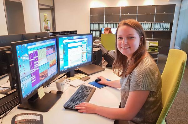 Jemand arbeitet am Schreibtisch an zwei Bildschirmen und blickt freundlich in die Kamera.