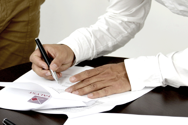 Jemand unterschreibt ein Dokument am Tisch, daneben liegen ein weiteres Blatt und ein Umschlag bereit.