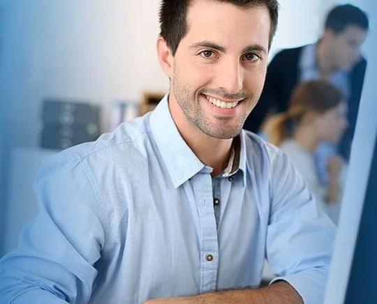 Ein Mitarbeiter lacht am Schreibtisch, während im Büro Kollegen im Hintergrund an ihren Aufgaben arbeiten.