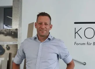 Ein Mitarbeiter steht im Ausstellungsraum neben einer Wand mit dem KOCH Forum für Bad & Wärme-Logo und berät Kundschaft.