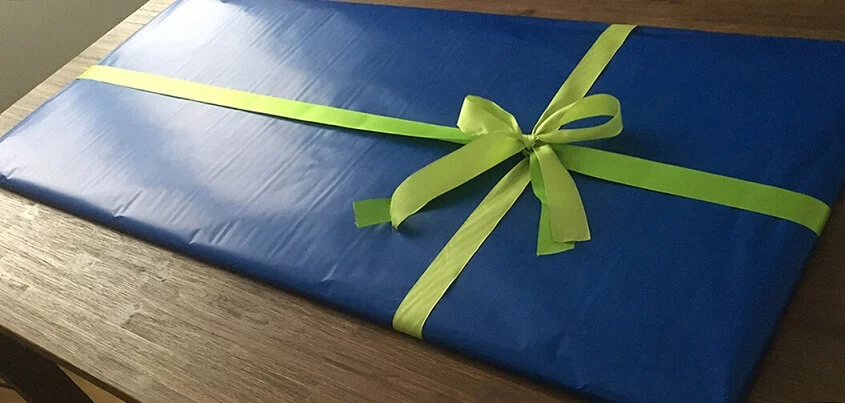 Zwei Hände legen ein in blauem Papier gewickeltes Geschenk mit grünem Band ordentlich auf einen Holztisch.